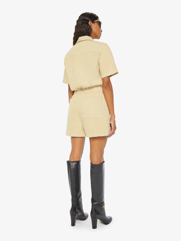 Back view of a woman in a light khaki  V-neck romper with a notched collar, short sleeves, patch pockets, buttons down the front and an elastic waistband. 