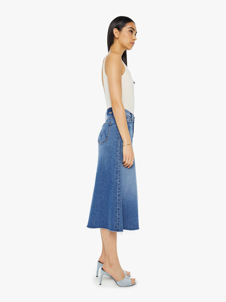 Side view of a woman high waisted midi skirt with an A-line fit and a frayed hem in medium blue wash.