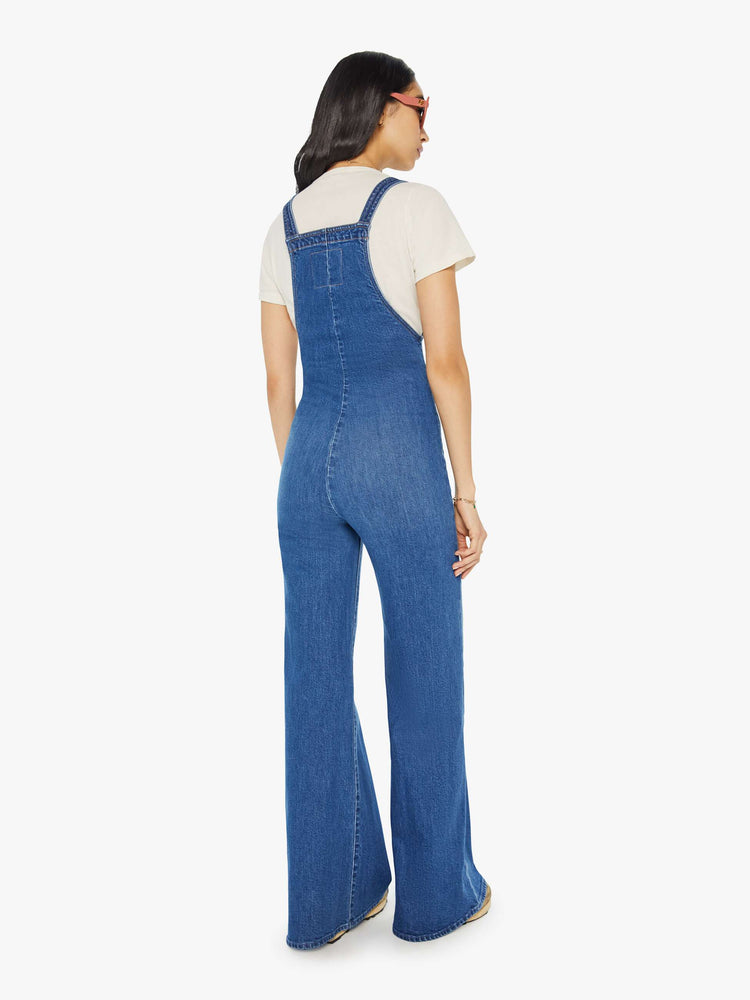 Back view of a woman in mid-blue denim overalls with a high rise, wide straight leg, patch pockets and a long 32-inseam with a clean hem.