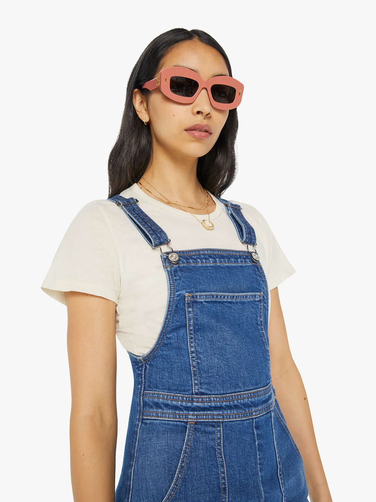 Detailed view of a woman in mid-blue denim overalls with a high rise, wide straight leg, patch pockets and a long 32-inseam with a clean hem.