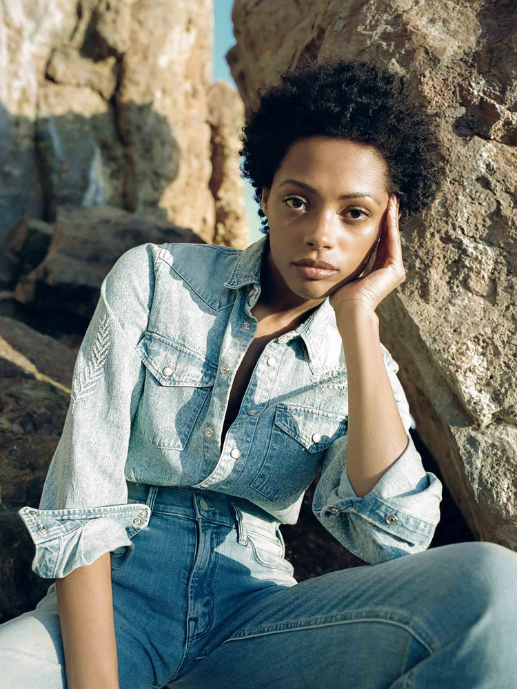 Editorial image of a woman sitting on large rocks, wearing a denim shirt and medium blue wash jeans.