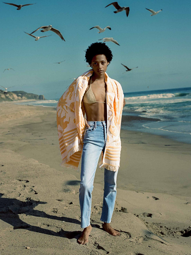 Editorial image of a woman standing on the beach, wearing a light blue wash jean and an orange patterned towel.