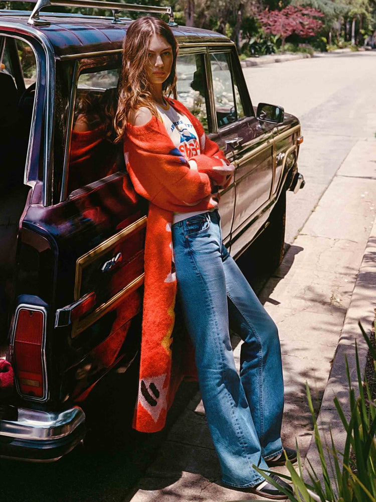 An editorial image of a woman leaning against a car, wearing medium blue wash jeans and a long red cardigan.