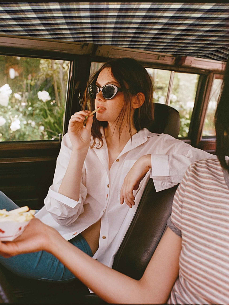 An editorial image of a woman in a car eating a french fry, wearing a white button down shirt and light blue pants.