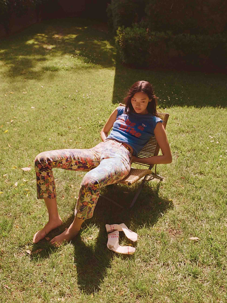 An editorial image of a woman sitting on a chair on the grass, wearing floral print pants and a blue graphic tee.