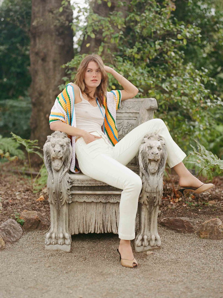 Editorial image of a woman sitting on a concrete hair in a garden, wearing cream pants and a knit shirt.