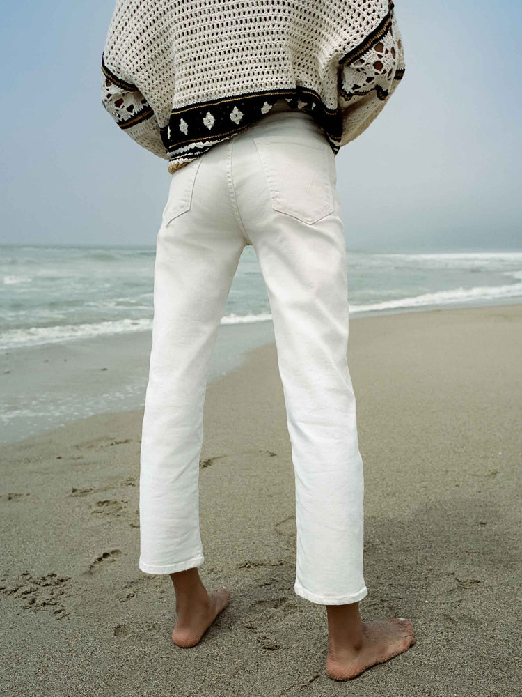 Editorial image of a woman facing the beach, wearing a cream and black knit sweater and cream wash jeans.
