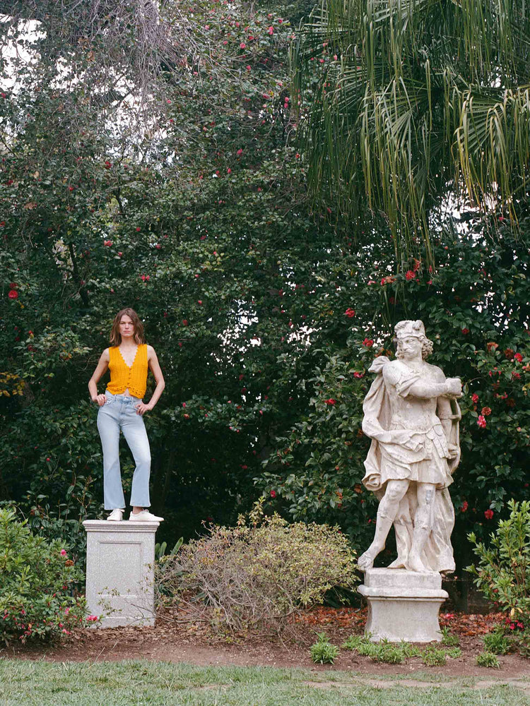 An editorial image of a woman standing on a pedestal, next to a statue, wearing a yellow knit vest and a light blue wash jean.