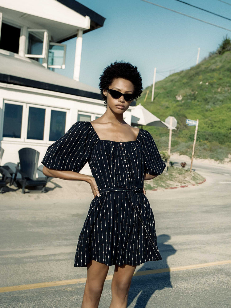 Editorial image of a woman wearing a black dress with white dots, standing in front of a house.