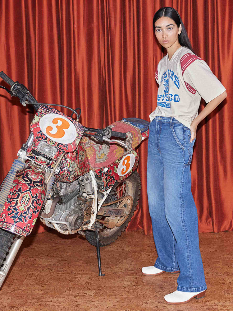 An editorial image of a woman standing next to a motorcycle, wearing medium blue wash denim carpenter jeans and a graphic tee.