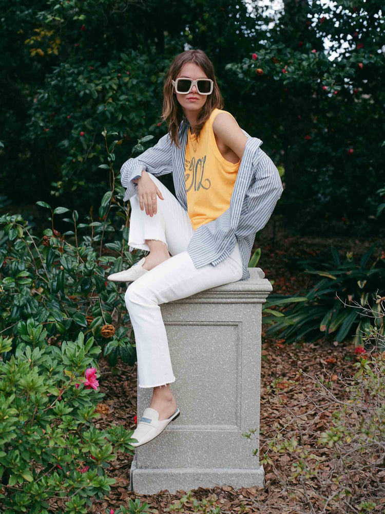 Editorial image of a woman sitting on a pedestal in a garden, wearing white jeans, a yellow tank, and a striped button down shirt.