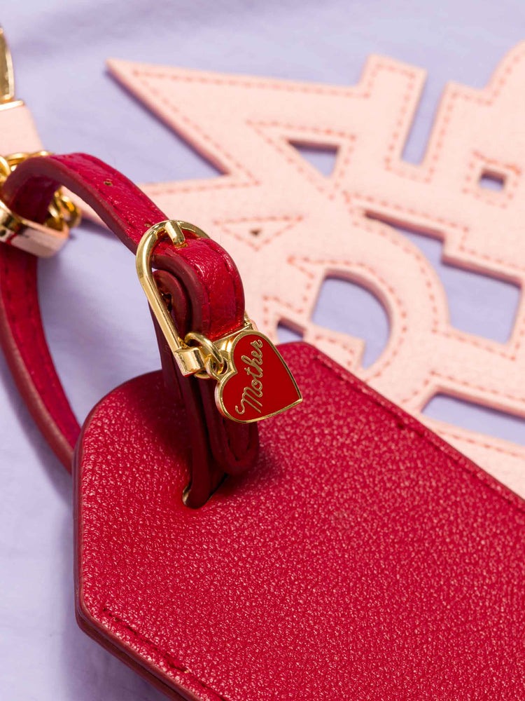 Close up view a luggage tag in burnt red faux leather with a clear plastic slot for your name and address, featuring a pink faux leather charm to remind you of MOTHER. 