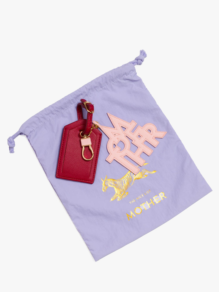 Flat  view of a luggage tag in burnt red faux leather with a clear plastic slot for your name and address, featuring a pink faux leather charm to remind you of MOTHER on top a purple bag.