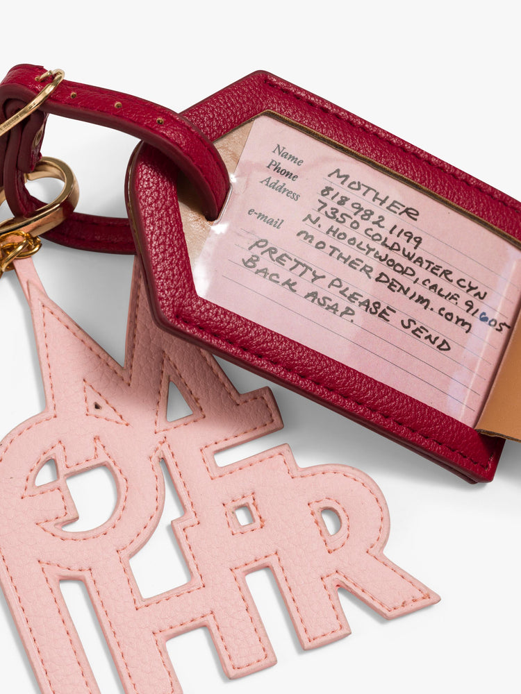 Close up view of a luggage tag in burnt red faux leather with a clear plastic slot for your name and address, featuring a pink faux leather charm to remind you of MOTHER. 