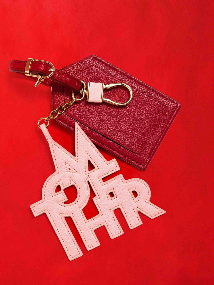 A luggage tag in burnt red faux leather with a clear plastic slot for your name and address, featuring a pink faux leather charm to remind you of MOTHER, against a red background. 