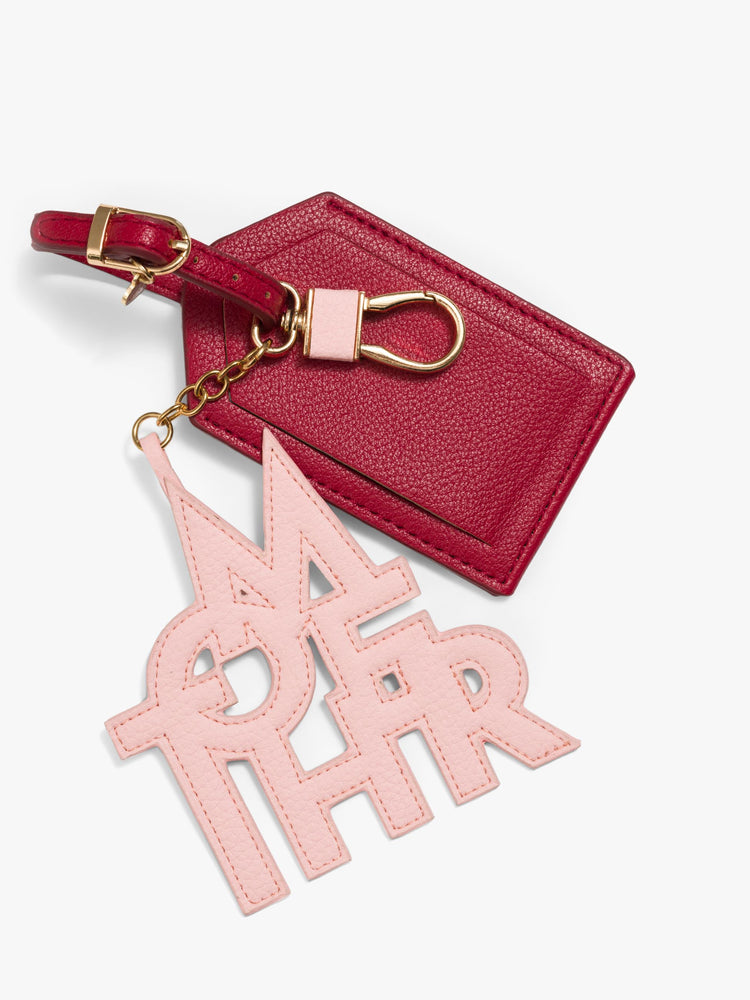 A luggage tag in burnt red faux leather with a clear plastic slot for your name and address, featuring a pink faux leather charm to remind you of MOTHER. 