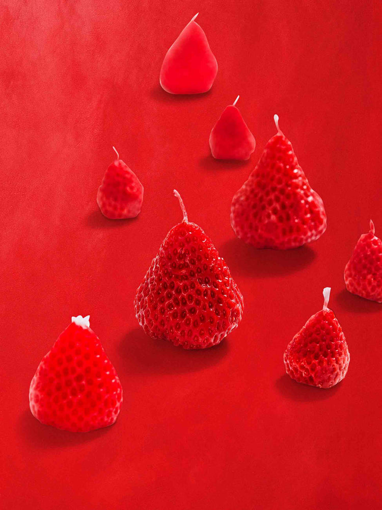 View of strawberry-shaped candle set made from a blend of soy wax and beeswax in light pink and red in a set of 8, against a red background.