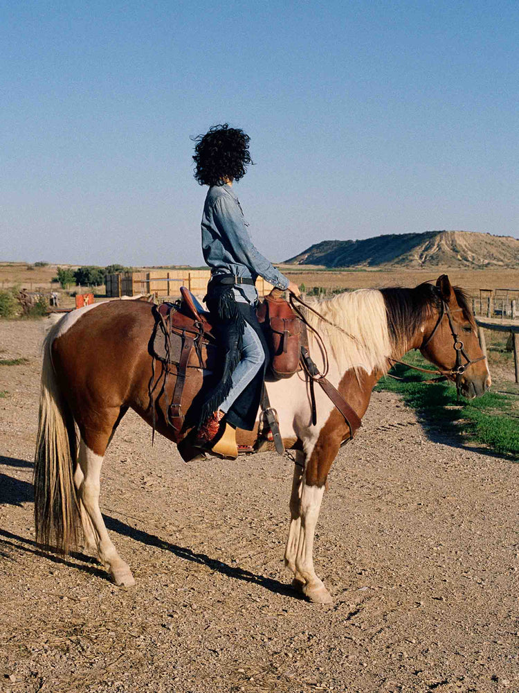 Editorial shot of a woman in denim button-up with front patch pockets and a curved hem in a light blue wash with snap buttons and metal tips on the collar on a horse