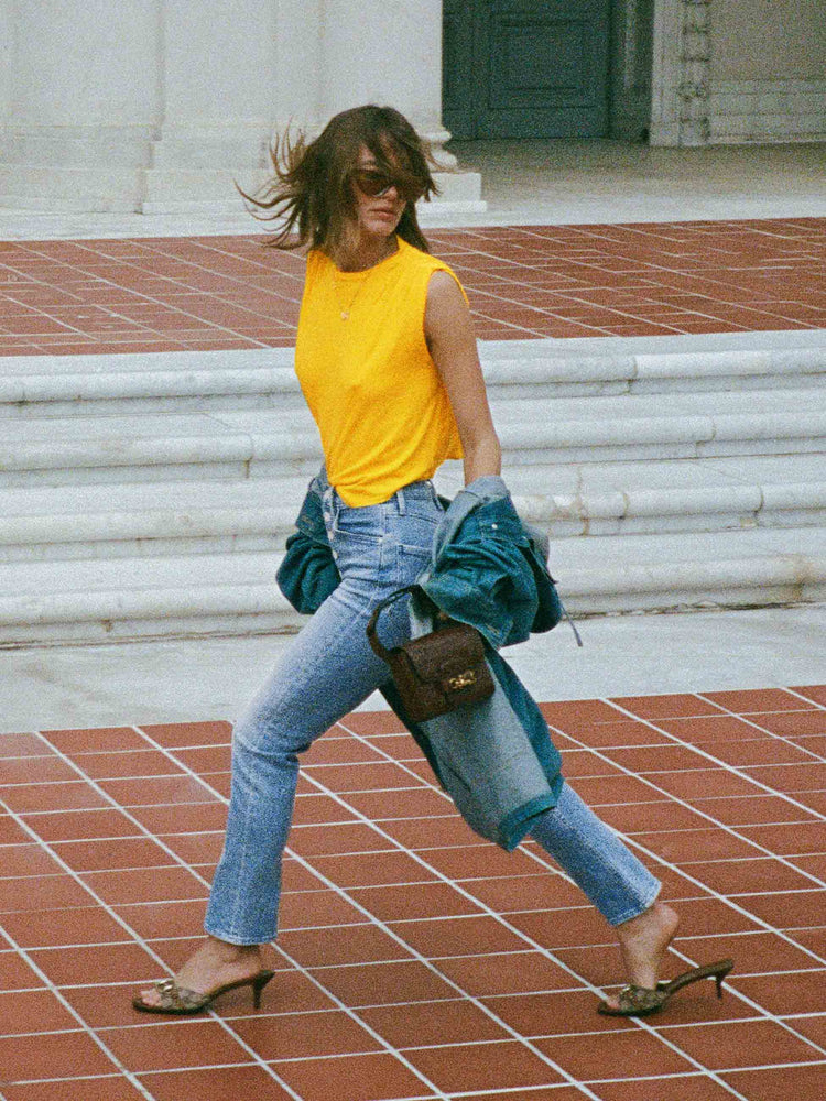 An editorial image of a woman walking outside a building, wearing a yellow tank top and medium blue wash jeans.