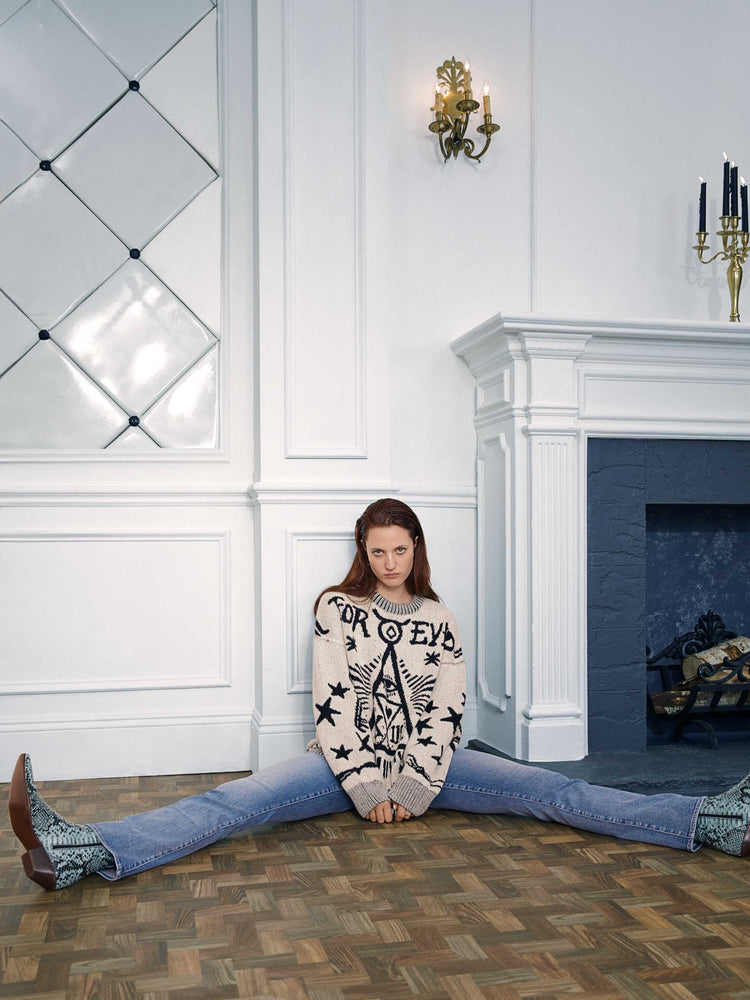 An editorial image of a woman sitting on the floor, wearing a cream sweater with large black graphics, paired with a medium blue wash jean.