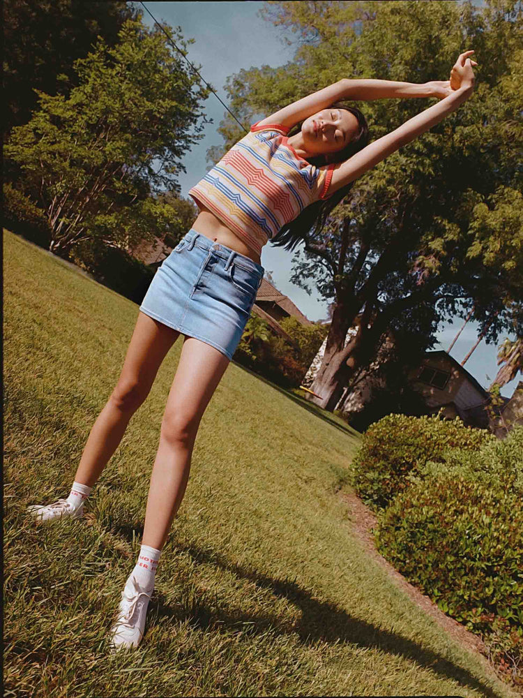 An editorial image of a woman standing on grass, stretching wearing a light blue wash denim skirt and a striped ringer tee.