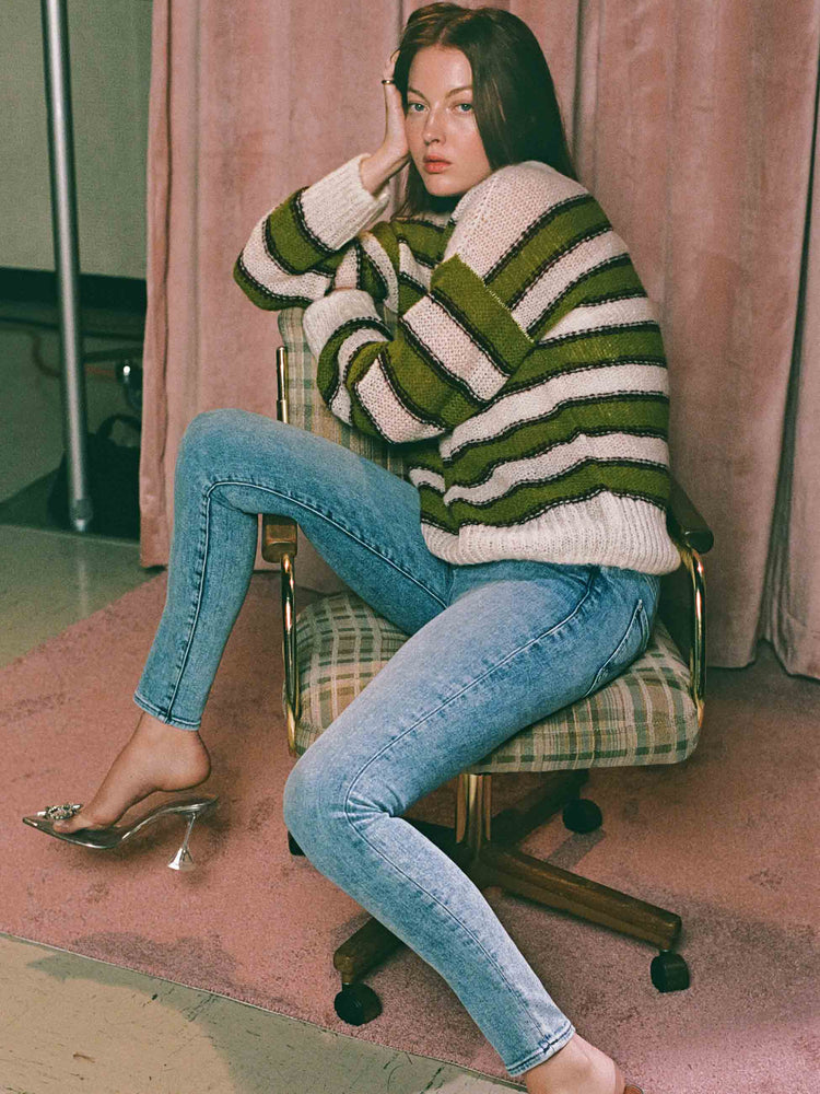 Editorial image of a woman sitting on a chair, wearing medium blue wash skinny jeans and a green and white striped sweater.