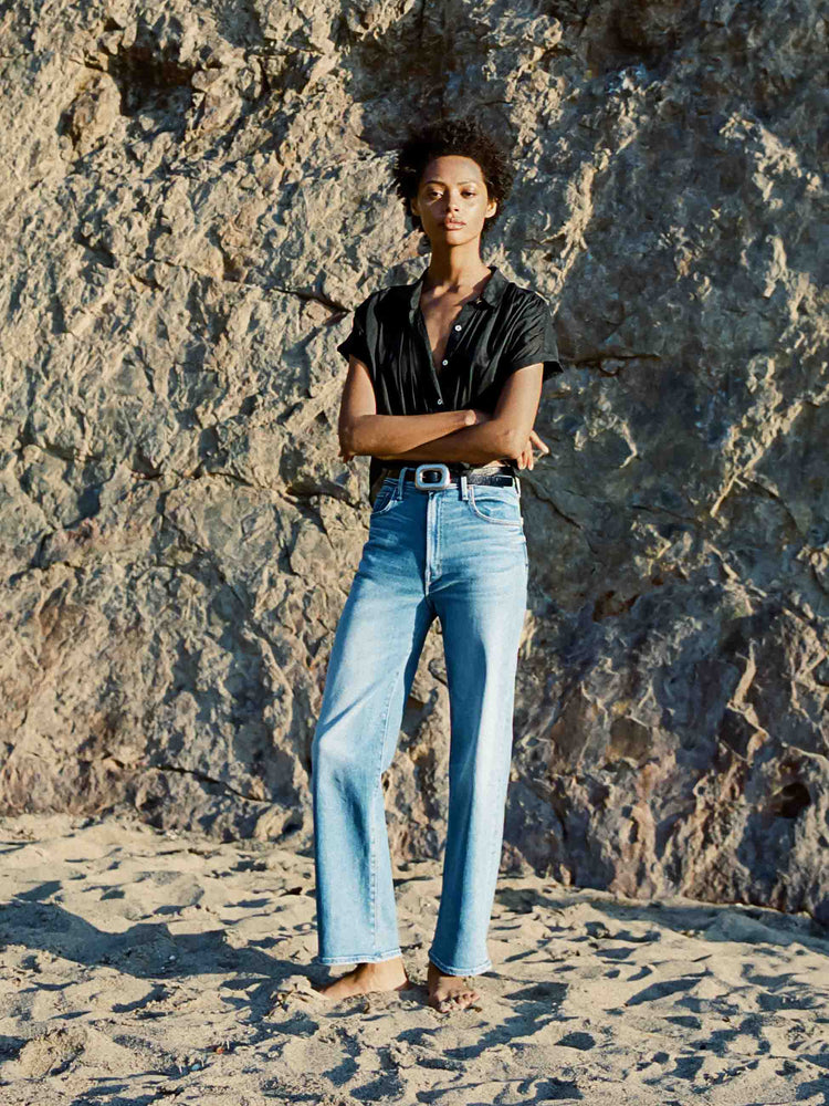 Editorial image of a woman standing in front of a stone wall on the beach, wearing a black button down top with short sleeves and a medium blue wash jeans.