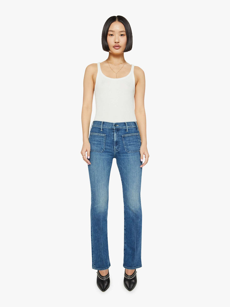 Front view of a woman in petite's medium blue high-rise bootcut designed with front patch pockets, a long inseam and a clean hem.