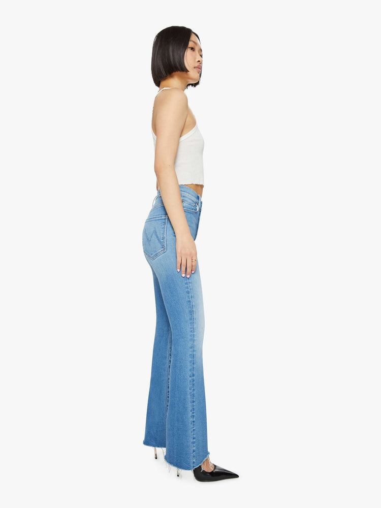 Side view of a petite woman in medium blue flare  jean has a high rise with a 29-inch length inseam and a frayed hem. 