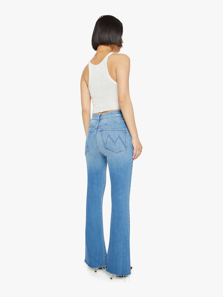 Back view of a petite woman in medium blue flare  jean has a high rise with a 29-inch length inseam and a frayed hem. 