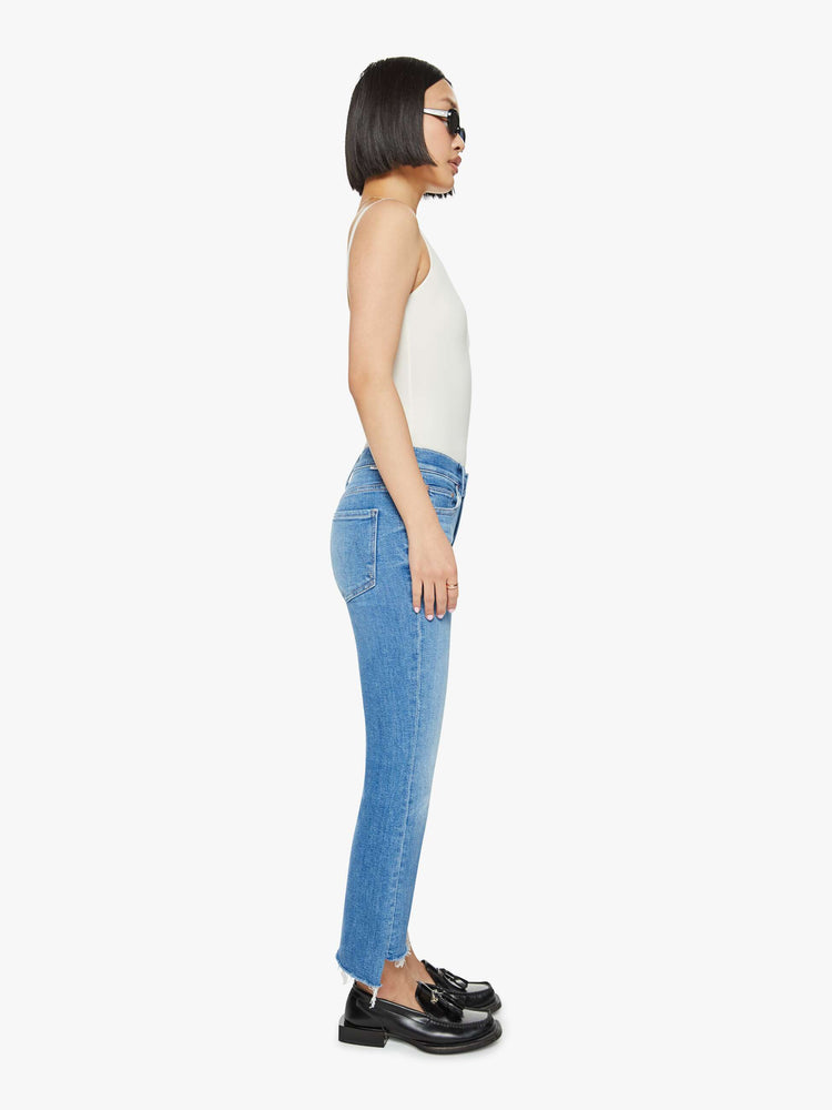 Side view of a petites woman high-waisted bootcut is cropped at the ankle with a frayed step-hem in a med blue wash.