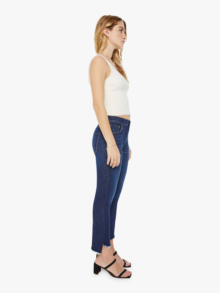 Side view of a petite woman in a dark blue wash high-waisted bootcut is cropped at the ankle with a frayed step-hem jean.