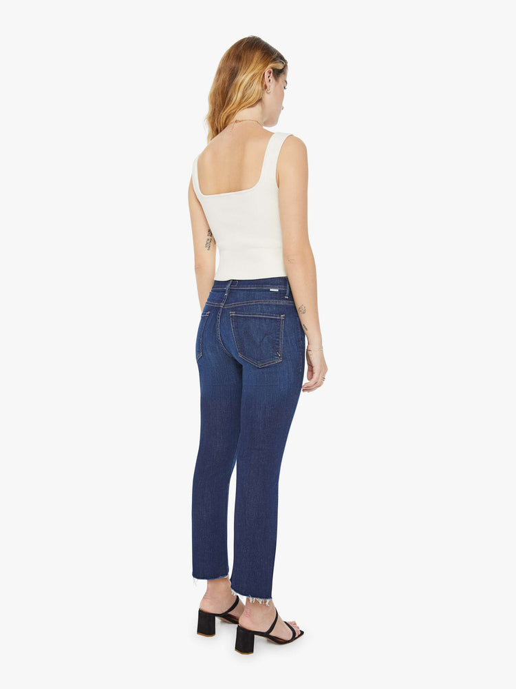 Back view of a petite woman in a dark blue wash high-waisted bootcut is cropped at the ankle with a frayed step-hem jean.