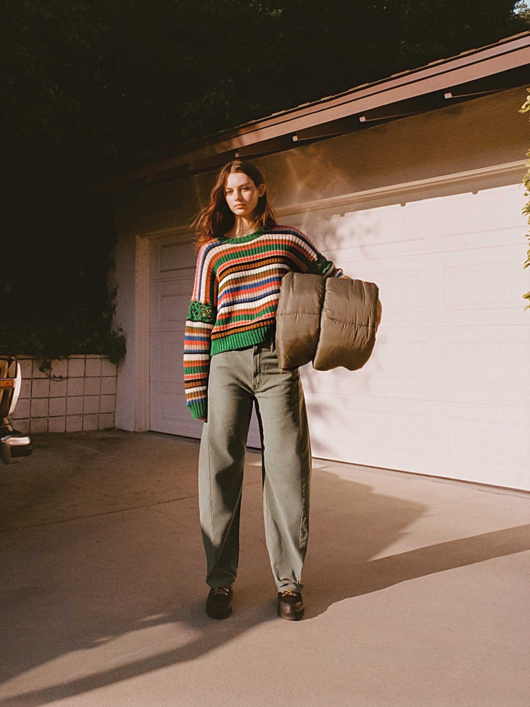 An editorial image of a woman standing in front of a garage holding a rolled sleeping bag, wearing army green pants and a striped knit sweater.