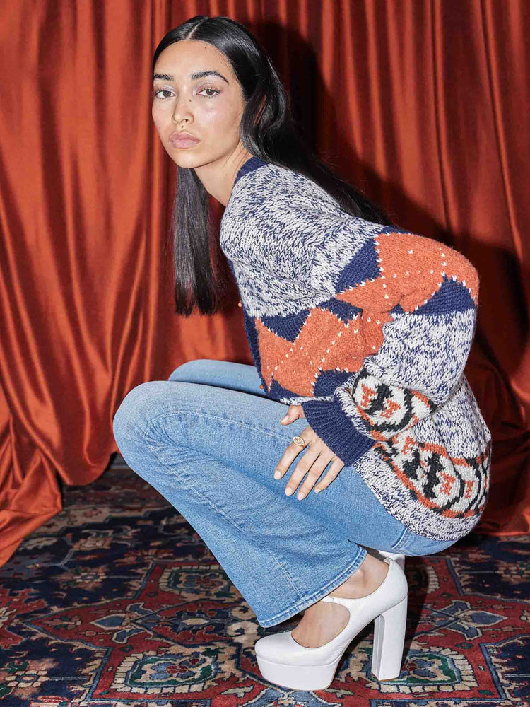 An editorial image of a woman squatting, wearing medium blue wash flare jeans and a patterned knit cardigan.