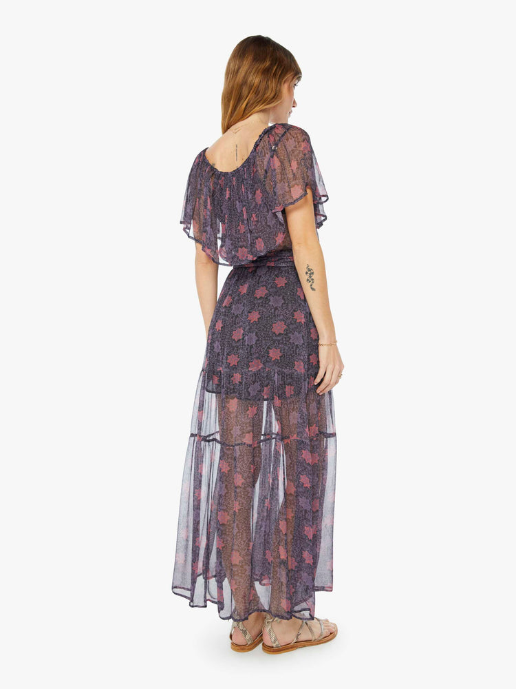 Back view of a woman sheer purple fabric with red floral print maxi dress with off-the-shoulder elastic neckline with an oversized ruffle, an optional tied waist and a tiered ankle-length skirt.