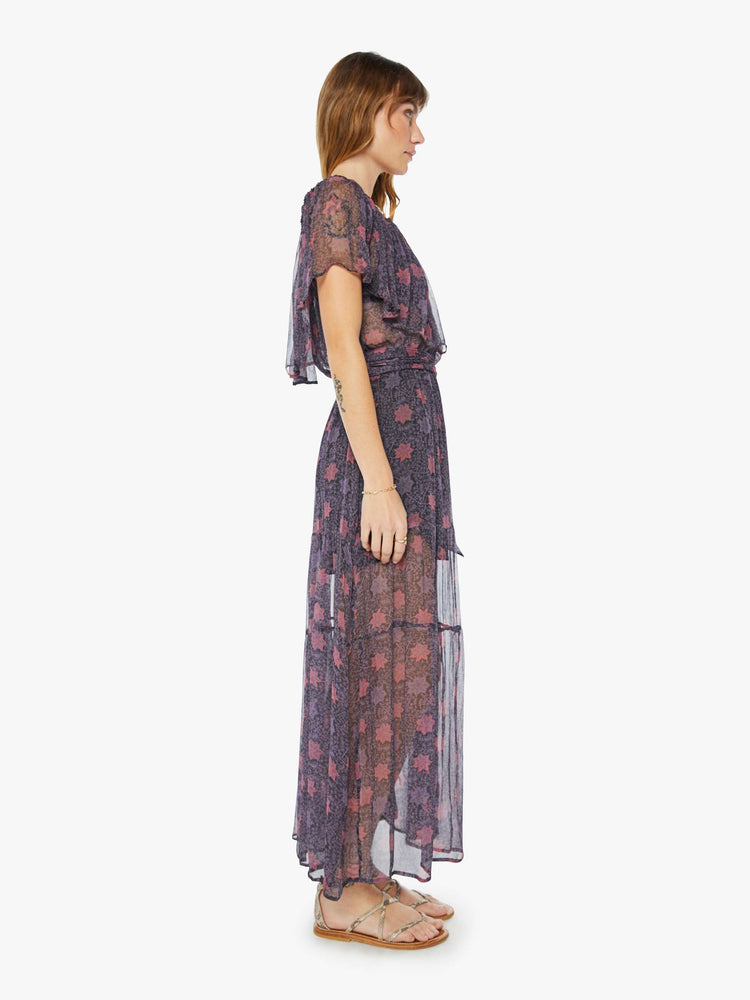 Side view of a woman sheer purple fabric with red floral print maxi dress with off-the-shoulder elastic neckline with an oversized ruffle, an optional tied waist and a tiered ankle-length skirt.