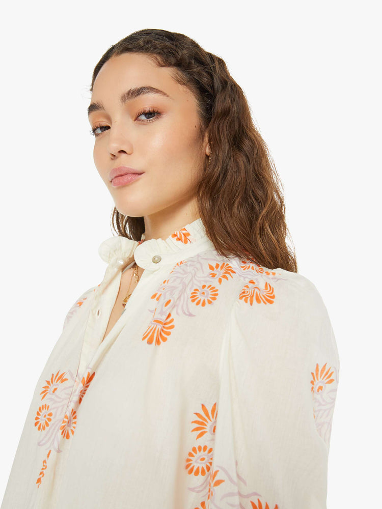 Close up view of a woman long sleeve blouse in white with a warm-toned tropical print, and features ruffled collar, puffed shoulders, long balloon sleeves, buttons down the front and a slightly cropped fit.