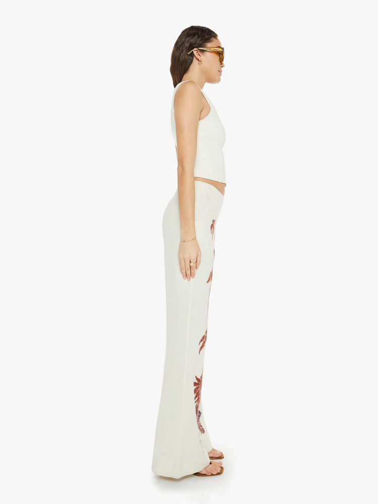 Side view of a woman high rise, slit pockets, a loose flared leg and slightly cropped hem in off-white with a floral design in maroon down the front of the legs.