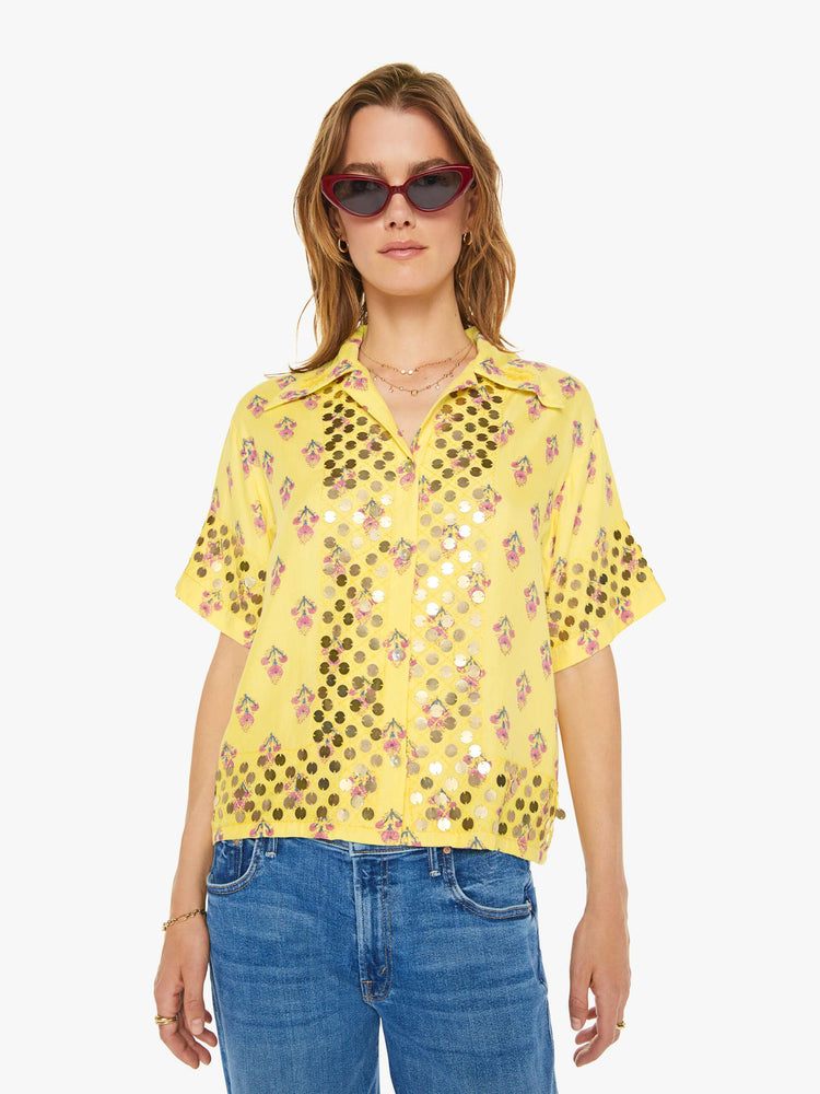 Front view of a woman short sleeve button-up in pastel yellow with a delicate floral print, and features a buttoned V-neck, slightly dropped shoulders, a boxy fit and coin embellishments throughout.