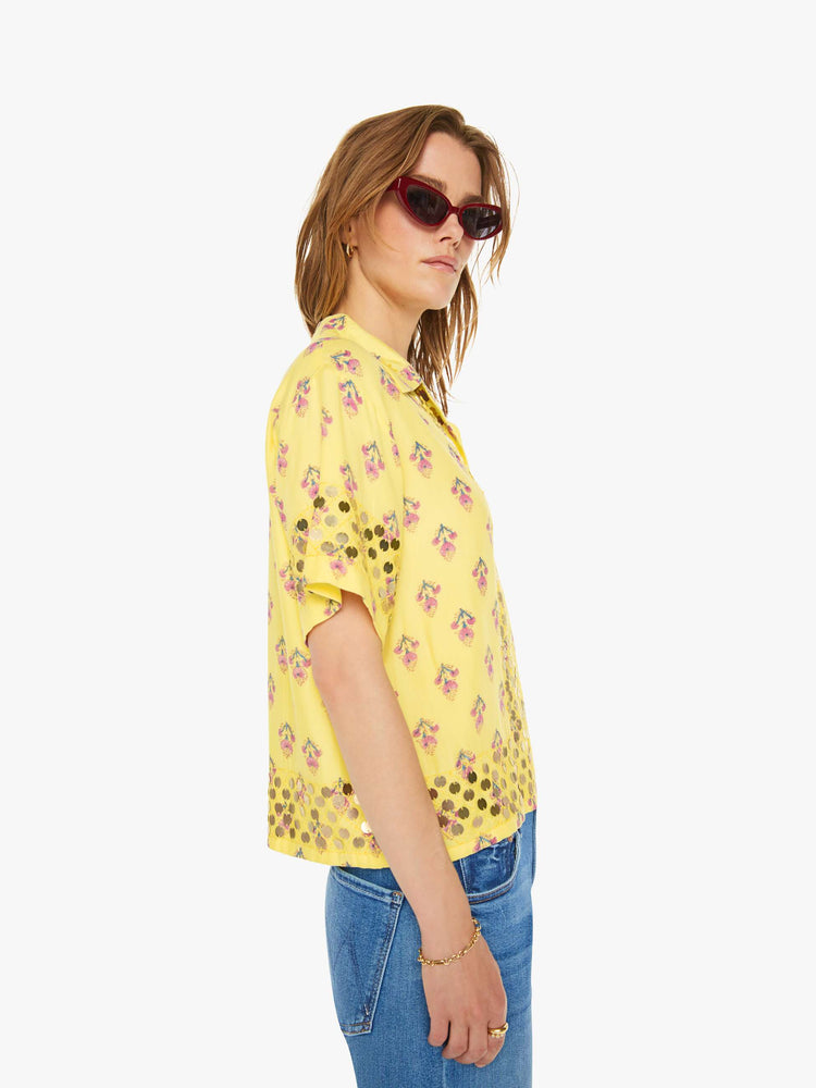 Side  view of a woman short sleeve button-up in pastel yellow with a delicate floral print, and features a buttoned V-neck, slightly dropped shoulders, a boxy fit and coin embellishments throughout.