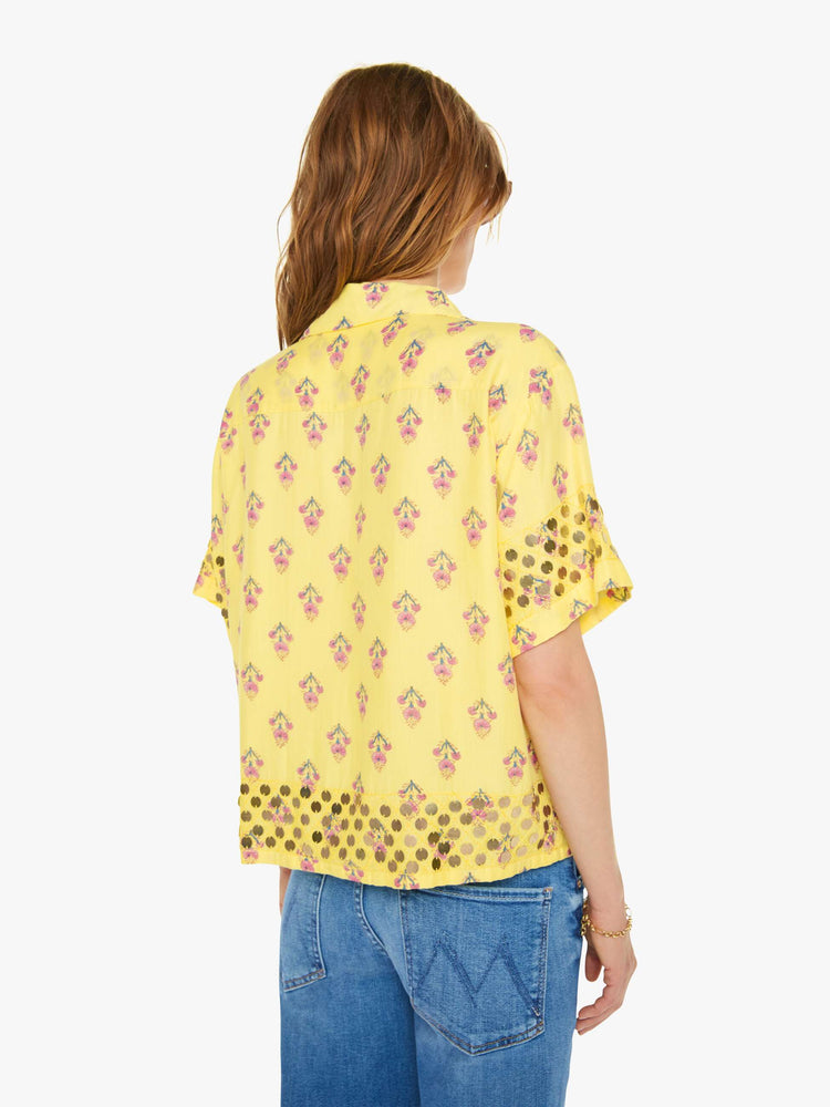 Back  view of a woman short sleeve button-up in pastel yellow with a delicate floral print, and features a buttoned V-neck, slightly dropped shoulders, a boxy fit and coin embellishments throughout.