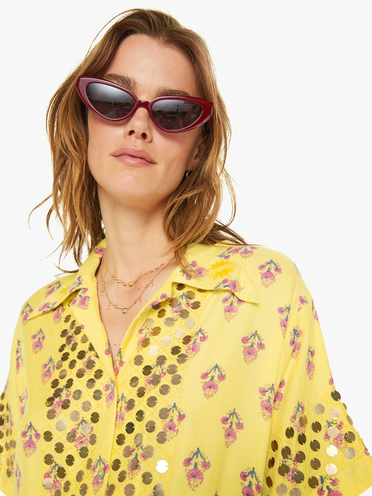 Close up  view of a woman short sleeve button-up in pastel yellow with a delicate floral print, and features a buttoned V-neck, slightly dropped shoulders, a boxy fit and coin embellishments throughout.