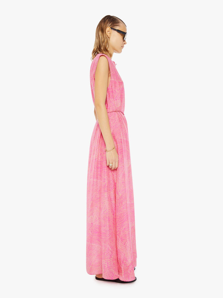 Side  view of a woman in pink with a delicate floral print, the sleeveless dress features a buttoned V-neck, a tied waist and a loose skirt with an ankle-grazing hem.