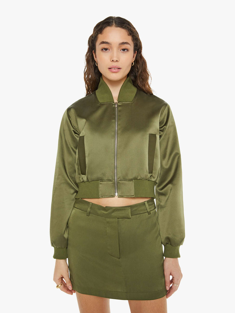 Front view of a woman in a green bomber jacker with a stacked collar, a zip closure, side slit pockets and a shrunken fit.