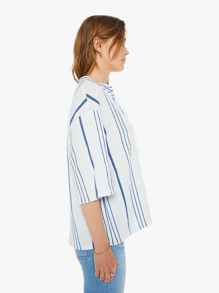 Side view of a woman in a white and blue striped blouse  designed with a V-neck that laces and ties, drop shoulders, elbow length sleeves and a loose, boxy fit. 