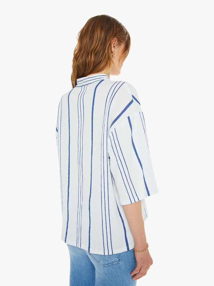 Back view of a woman in a white and blue striped blouse  designed with a V-neck that laces and ties, drop shoulders, elbow length sleeves and a loose, boxy fit. 