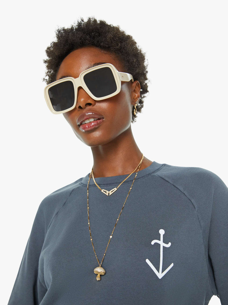 Close up view of a woman in a dark grey sweatshirt with a relaxed shape with raglan sleeves and is decorated with a white anchor graphic on the chest.