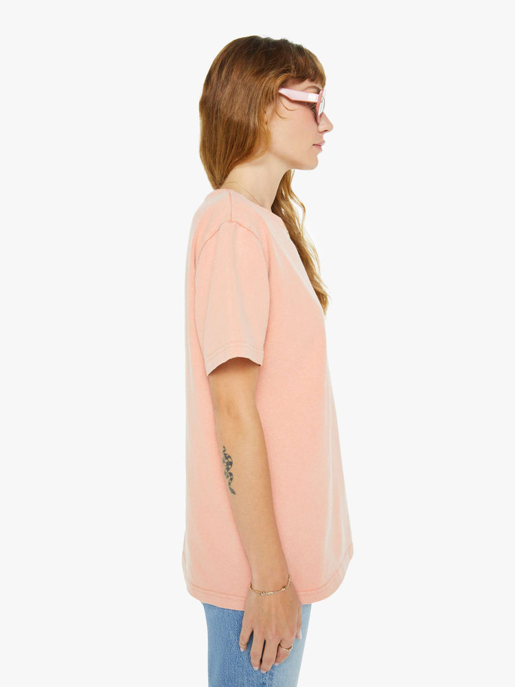 Side view of a woman light peach tee with a white anchor graphic on the chest, has a ribbed crewneck and a straight fit.