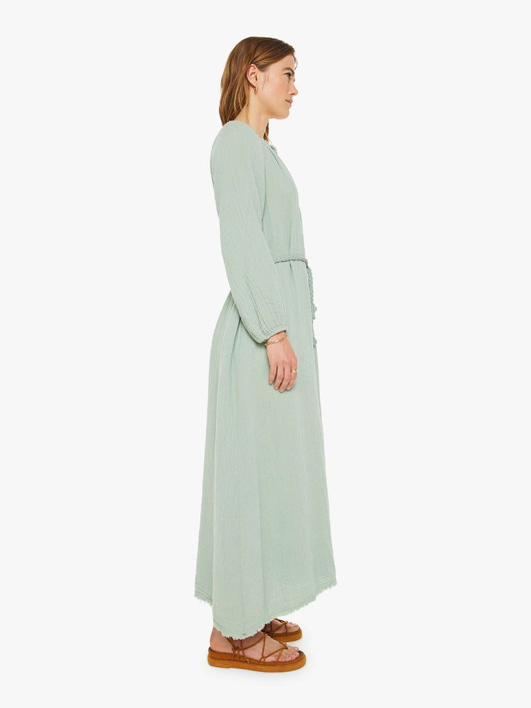 Side view of a woman maxi dress designed with a V-neck, long balloon sleeves, buttons down the front, a tied waist and ankle-grazing hem in a soft green gray.